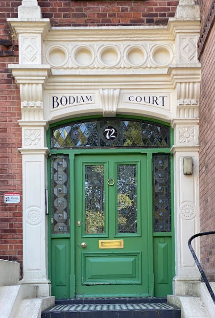 A detailed view of a residential building entrance displaying a bright green wooden front door with two glass panels featuring decorative diamond-shaped leaded glass designs, situated within a cream-colored stone surround with ornate carvings and geometric patterns. Above the door, a semi-circular stained glass window is visible, surrounded by a stone arch with the address '72' in black numerals. To the left of the door, a vertical white metal handle is mounted on the door, and below the handle, a letterbox slot is integrated into the door. The entrance is set against a red brick facade, and to the right, a black metal handrail is partially visible, leading down a small staircase with stone steps. The top of the image shows part of another brick wall, and the overall environment suggests the building is part of an urban residential complex. This setting is typical for a property undergoing home relocation or furniture transport, with an emphasis on the detailed architectural features of the door and doorway.