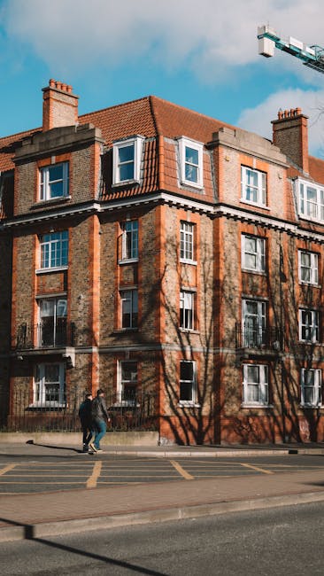 A multi-storey red-brick building with a sloped terracotta tiled roof, featuring multiple white-framed windows, some with open casements. The building has stone or concrete detailing around the windows and edges, and chimneys on the roof. In front of the building, two individuals are walking along the pavement; one appears to be carrying a bag. To the right, a large construction crane extends into the sky, indicating nearby construction activity. The foreground shows part of a street with yellow line markings along the curb, and the image is illuminated by natural daylight with a partly cloudy sky in the background. The scene depicts an urban environment consistent with a residential area, relevant to moving and home relocation services offered by Man and Van Wood Green, especially in the context of packing, furniture transport, and loading process in the N22 postcode area.